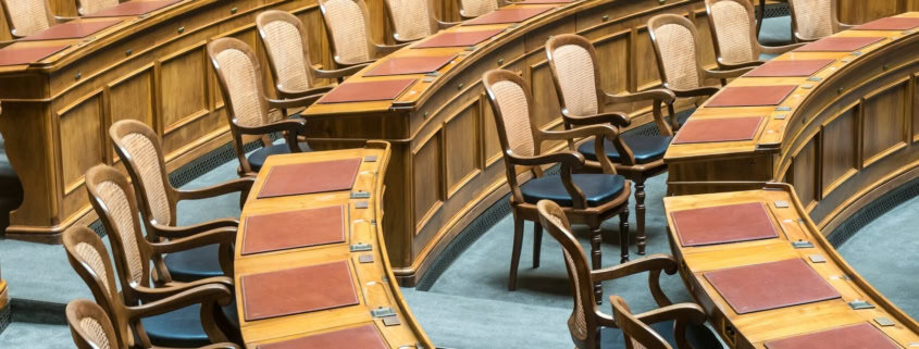 Rows of wooden chairs with cane backs and leather desks in a legislative chamber
